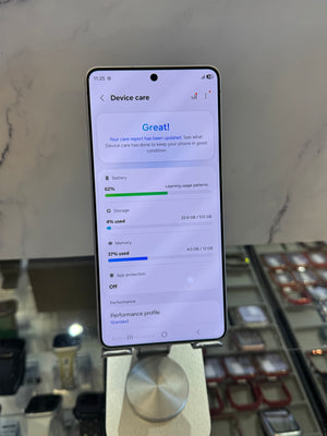 A smartphone on a metal display stand showing the device care screen with battery and storage usage details, photographed in a retail or showroom setting with display cases in the background.