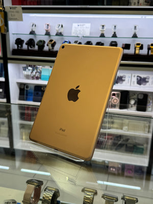 A gold-colored iPad shown from the back, resting on a clear glass display counter inside a retail store. The Apple logo is clearly visible in the center, with blurred watch displays in the background.