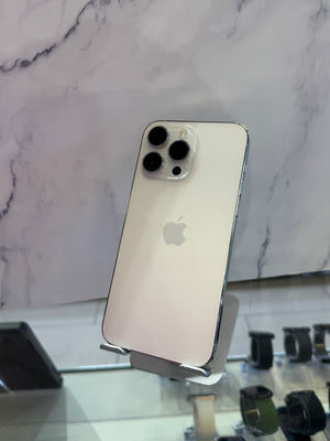A silver Apple iPhone 12 Pro shown from the back, resting on a clear display stand in front of a white marble-patterned wall.