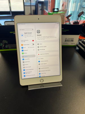 A white tablet device displayed upright on a stand on a dark tabletop, with a blurred store or office background and a visible Apple logo on the screen interface.