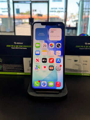 A black smartphone with a lit home screen displayed upright on a dark charging stand, with retail packaging boxes behind it and a windowed wall in the background.