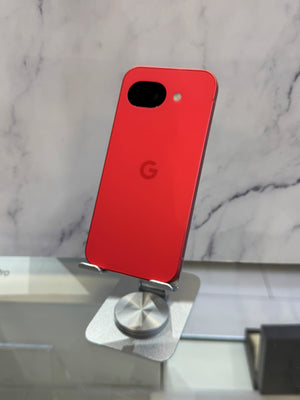 A red Google Pixel smartphone shown from the back, resting on a small metallic display stand against a light marble-patterned background.