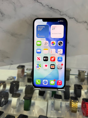 A black iPhone with its screen turned on, displaying the home screen and widgets, resting on a white marble-patterned surface with a blurred watch display case in the background.