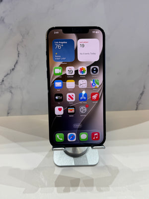 A black iPhone with a cracked screen protector, displayed upright on a silver metal stand against a white marble-patterned background.