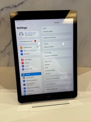 An iPad standing upright on a clear display stand, showing the Settings menu on screen against a light marble-patterned background.