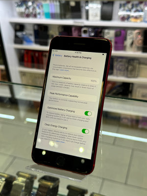 A red smartphone displaying an iOS battery health and charging settings screen, standing on a clear display stand inside a retail store with blurred shelves in the background.