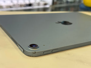 A close-up shot of the back of a gray Apple iPad resting on a light beige surface, showing the camera lens, side buttons, and part of the Apple logo.