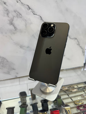 A dark gray Apple iPhone 12 Pro displayed on a silver stand, showing its rear camera array and the Apple logo, set against a white marble wall background.