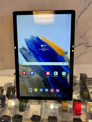 A Samsung Galaxy tablet with a black bezel and a colorful abstract wallpaper, displayed on a clear acrylic stand in a retail setting with other smartwatches in the background.