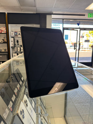 A black tablet device with a home button, photographed standing upright on a glass display counter inside a retail store. Other electronics are visible in the background.