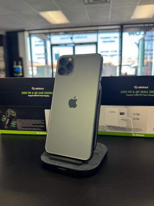 A silver Apple iPhone 12 Pro shown from the back, resting on a gray stand in front of its black and green retail box on a dark tabletop.