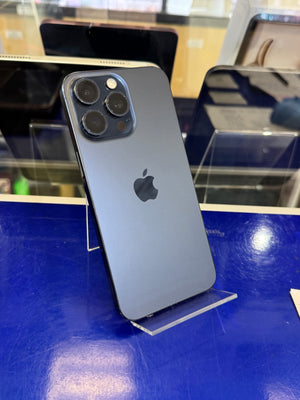 A dark blue-gray Apple iPhone Pro shown from the back, standing on a clear acrylic display stand on a blue counter inside a store.