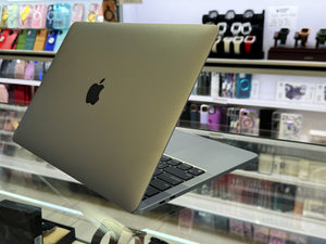 A silver Apple MacBook laptop with a black Apple logo, shown open and angled on a glass display counter inside a retail store.
