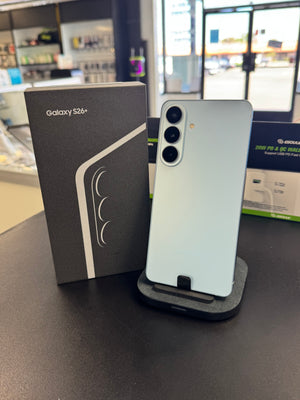 A Samsung Galaxy S26+ smartphone in a pale icy blue-green color stands on a black charging dock on a dark tabletop, next to its original black retail box. The background shows a blurred retail store environment.