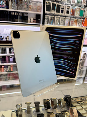 A gold Apple iPad shown from the back, with its screen turned on and displaying a colorful wallpaper. The iPad is photographed inside a retail store with shelves of other electronics in the background.
