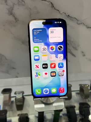 A black Apple iPhone with a lit screen displaying the home screen, resting on a silver display stand on a white and gray marble surface.
