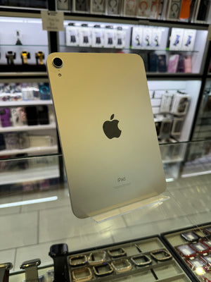 A gold Apple iPad shown from the back, standing on a clear display stand inside a retail store with shelves of accessories in the background.