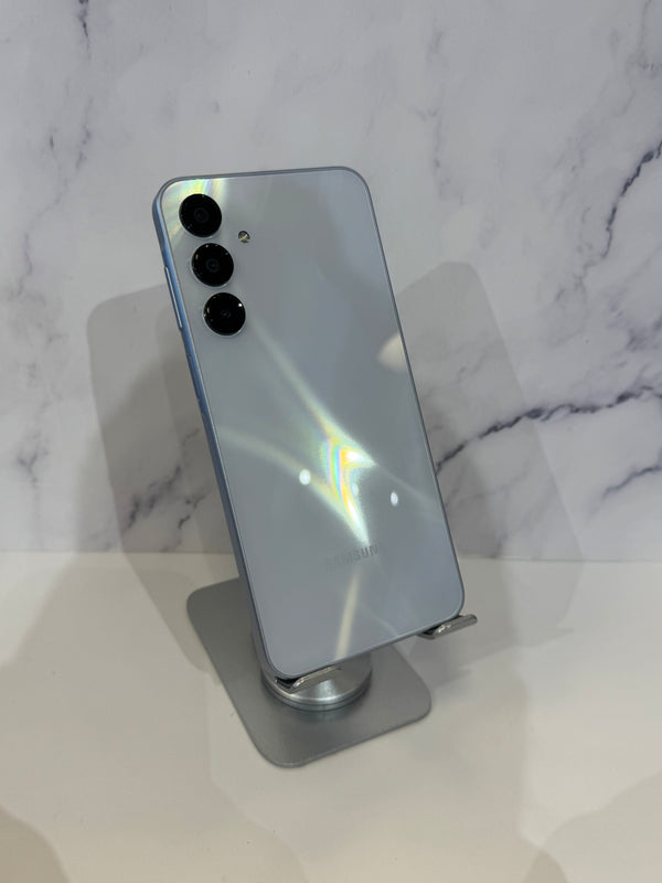A light blue-gray Samsung Galaxy smartphone shown from the back, resting on a silver metal display stand against a white marble-patterned background.