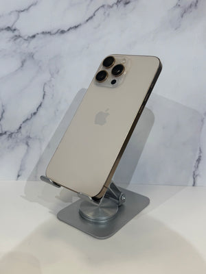An Apple iPhone with a light gold backplate and dark gray camera lenses, displayed on a silver metal stand against a white and gray marble-patterned background.