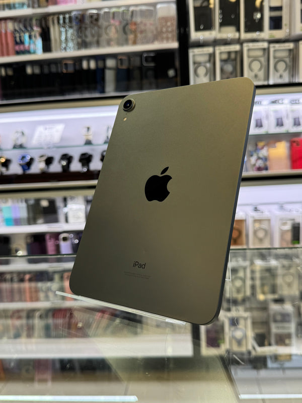 A grey-colored iPad shown from the back, resting on a glass display counter inside a retail store with shelves of accessories in the background.