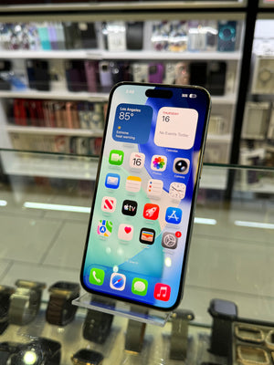 A black iPhone with a lit home screen displaying various colorful apps, resting on a clear acrylic display stand inside a retail store.