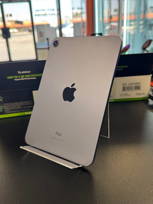 A silver Apple iPad shown from the back, standing on a clear display stand on a dark tabletop. The Apple logo is visible in the center, with retail packaging boxes in the blurred background.