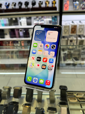 A black smartphone with a lit home screen, standing on an acrylic stand in a retail display with blurred store fixtures in the background.