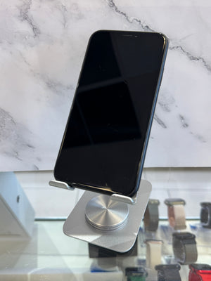 A black smartphone with a blank screen stands upright on a silver metal display stand. The phone is photographed indoors against a white marble-patterned wall, with blurred watch displays visible below on a glass counter.