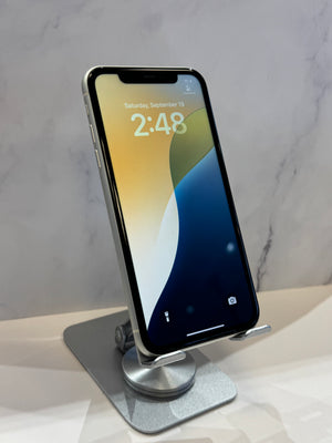A silver smartphone with a black front display stands upright on a metallic silver stand against a light gray marble-patterned background.