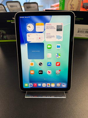 An iPad with a black bezel and silver front face, displaying its home screen on a clear acrylic stand on a dark tabletop. The background shows a retail or office environment with boxes and a baseball visible out of focus.