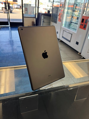 An Apple iPad with a metallic gold finish, showing the back of the device with the Apple logo. It is displayed on a clear acrylic stand on a glass counter inside a retail store.