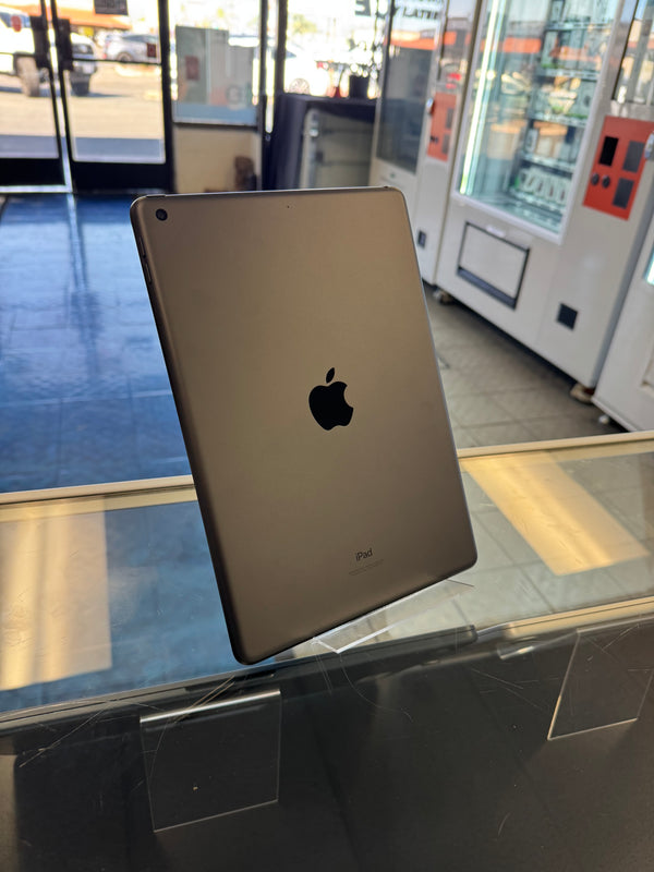 An Apple iPad with a metallic gold finish, showing the back of the device with the Apple logo. It is displayed on a clear acrylic stand on a glass counter inside a retail store.