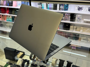 A silver Apple MacBook laptop is shown open on a display stand inside a retail store, with shelves of accessories blurred in the background.
