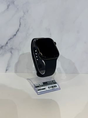 A black smartwatch with a rectangular black screen and black band, displayed on a small clear plastic stand against a white marble-patterned background.