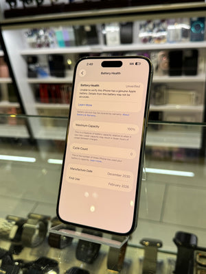 A black smartphone displayed upright in a glass retail case, with its screen on showing settings pages. The store interior with shelves and fixtures is visible through the glass in the background.