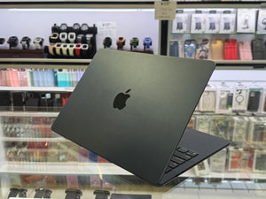 A dark gray Apple MacBook laptop is shown open on a glass display counter inside a retail store, with shelves of accessories and watches blurred in the background.