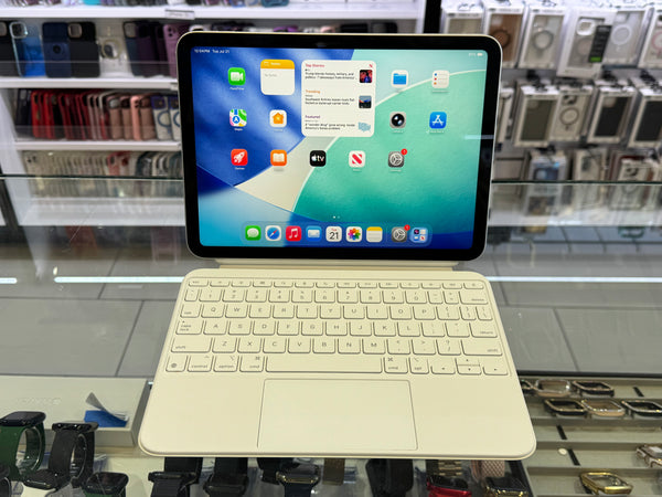 A white iPad with a matching keyboard cover and trackpad, displayed on a stand in a retail store. The screen is turned on and shows the iPad home screen.