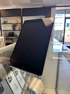 A black Apple iPad tablet displayed on a glass stand inside a retail store, with other devices and store fixtures visible in the background.