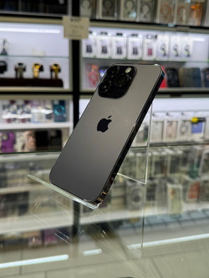 A dark gray Apple iPhone Pro displayed on a clear acrylic stand inside a retail store, with blurred shelves of phones and accessories in the background.
