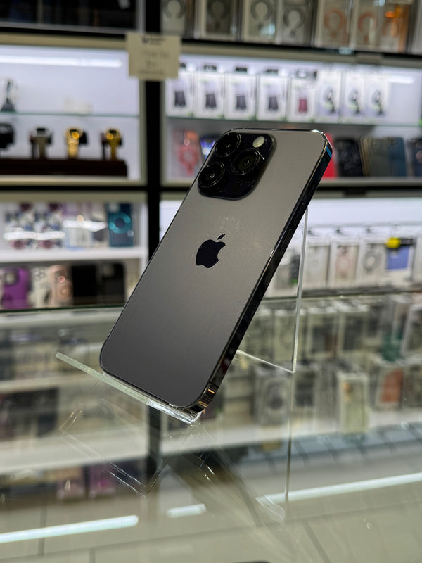 A dark gray Apple iPhone Pro displayed on a clear acrylic stand inside a retail store, with blurred shelves of phones and accessories in the background.