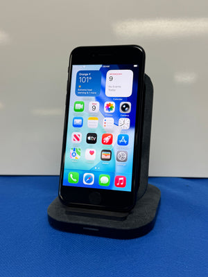 A black smartphone displayed upright on a dark charging dock against a light gray background, resting on a blue surface.