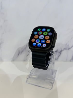 A black smartwatch with a rectangular rounded-corner display showing colorful app icons, mounted on a clear acrylic stand against a white marble-patterned background.