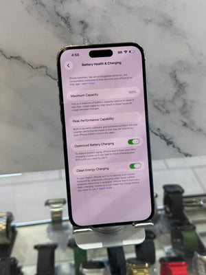 A black smartphone lying on its side on a white marble-like surface, displaying a settings screen with battery-related options. The background is a blurred retail or office environment with shelves and objects.