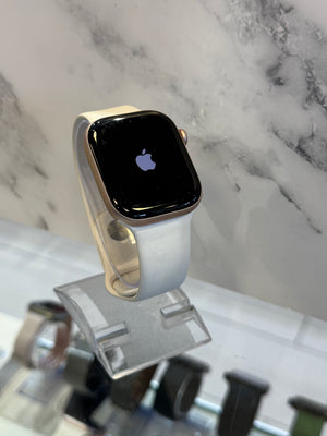 An Apple Watch with a light gray band and gold-toned case, displayed on a clear stand against a white marble-patterned background.