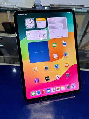 An iPad Pro tablet standing upright on a clear display stand, showing the home screen with colorful app icons and widgets. The device is photographed in a retail or showroom setting with a blue counter and glass display elements in the background.