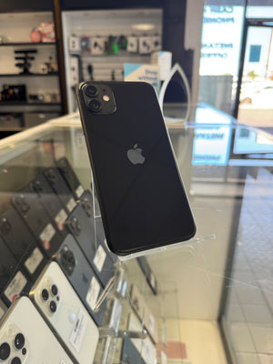 A black Apple iPhone shown from the back, standing on a clear display stand inside a retail store. The Apple logo is visible in the center, and other phones are displayed in the background through a glass case.