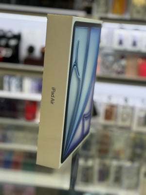 An Apple iPad Air box standing upright in a blurred retail store setting. The packaging is cream-colored with a blue and white graphic of the tablet on the front and 'iPad Air' printed vertically on the side.