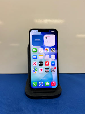 A black smartphone with its screen on, displaying the home screen, resting on a black charging dock against a light gray wall and blue surface.