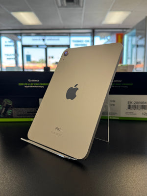 A silver Apple iPad shown from the back, standing on a clear acrylic display stand on a dark tabletop. The Apple logo and the word 'iPad' are visible on the back of the device.