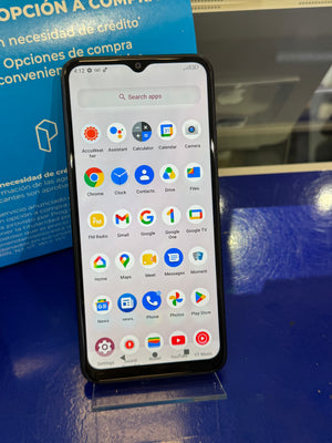 A black smartphone with a front-facing camera cutout stands on a clear display stand on a blue surface, showing the Android home screen with app icons. A blue promotional sign is partially visible in the background.